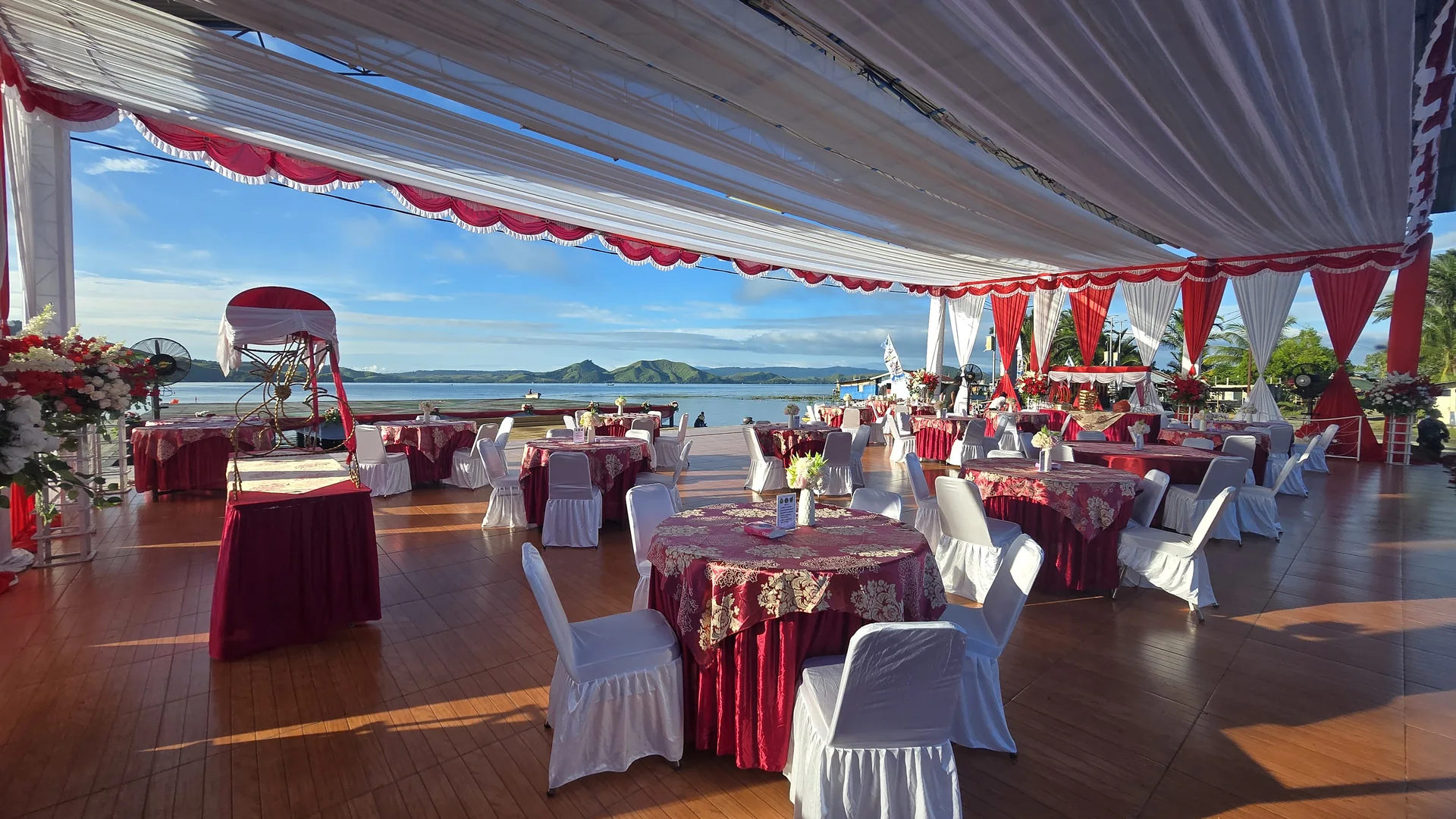 Tenda event outdoor dengan meja bundar, kursi, dekorasi merah-putih, dan pemandangan laut.